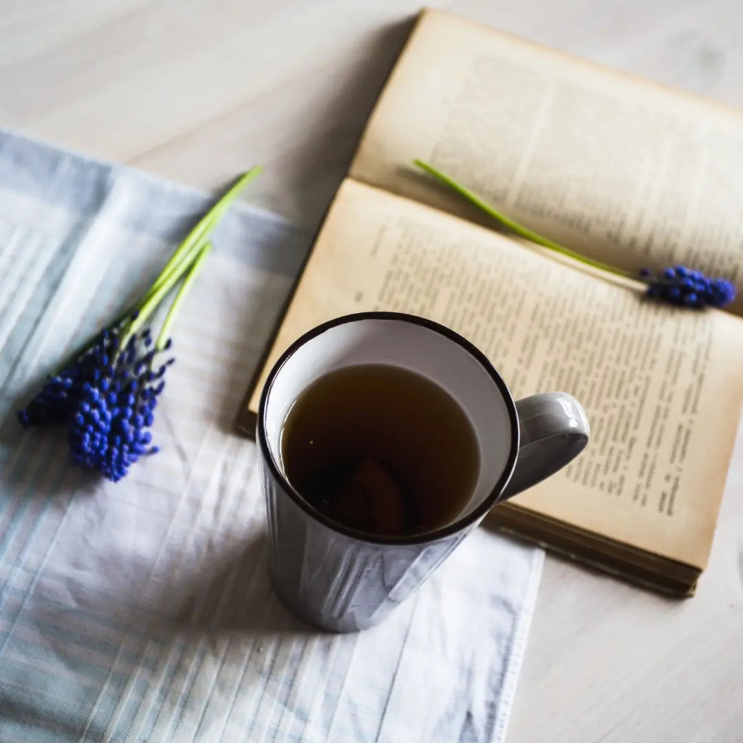 A cosy reading setup featuring a colourful hardback book and a steaming mug of Wander Cuppa fruit tea for a Beltane celebration.